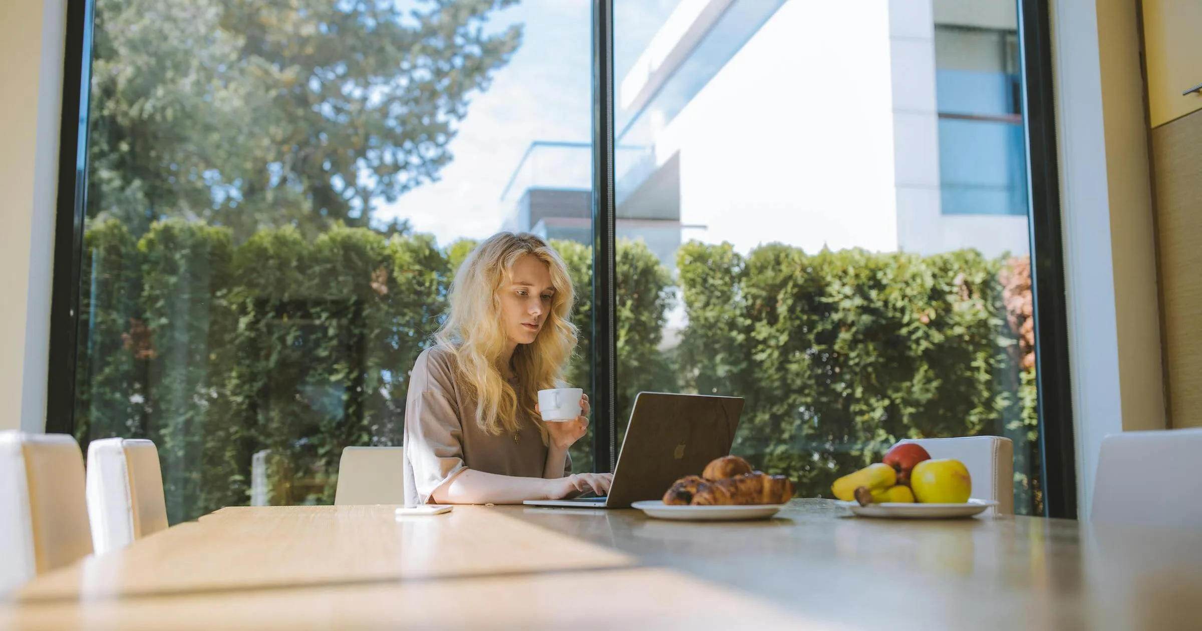 Woman reading on a laptop with coffee at a sunlit dining table